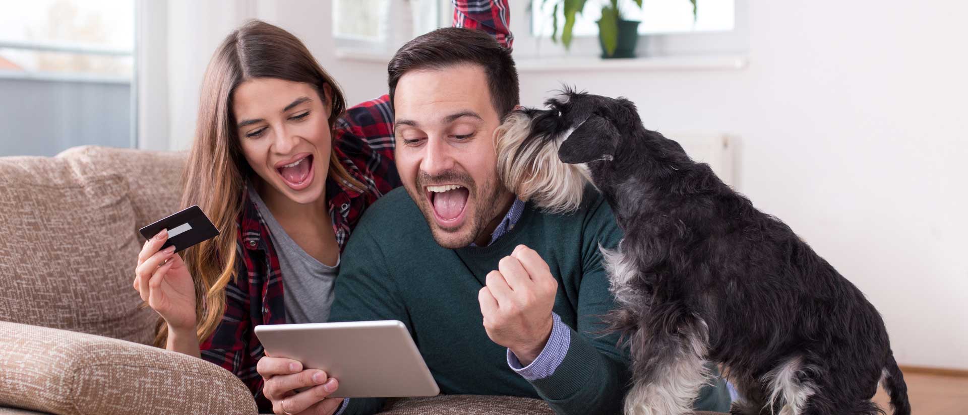 Couple and dog laying on the bed with their laptop and credit card in hand looking happy while buying something online.